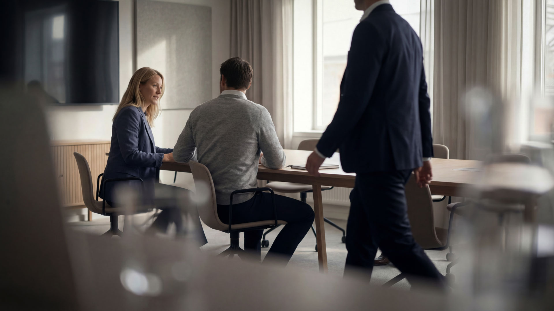 Zwei Personen sitzen an einem Konferentisch, eine weitere Person läuft durch das Bild