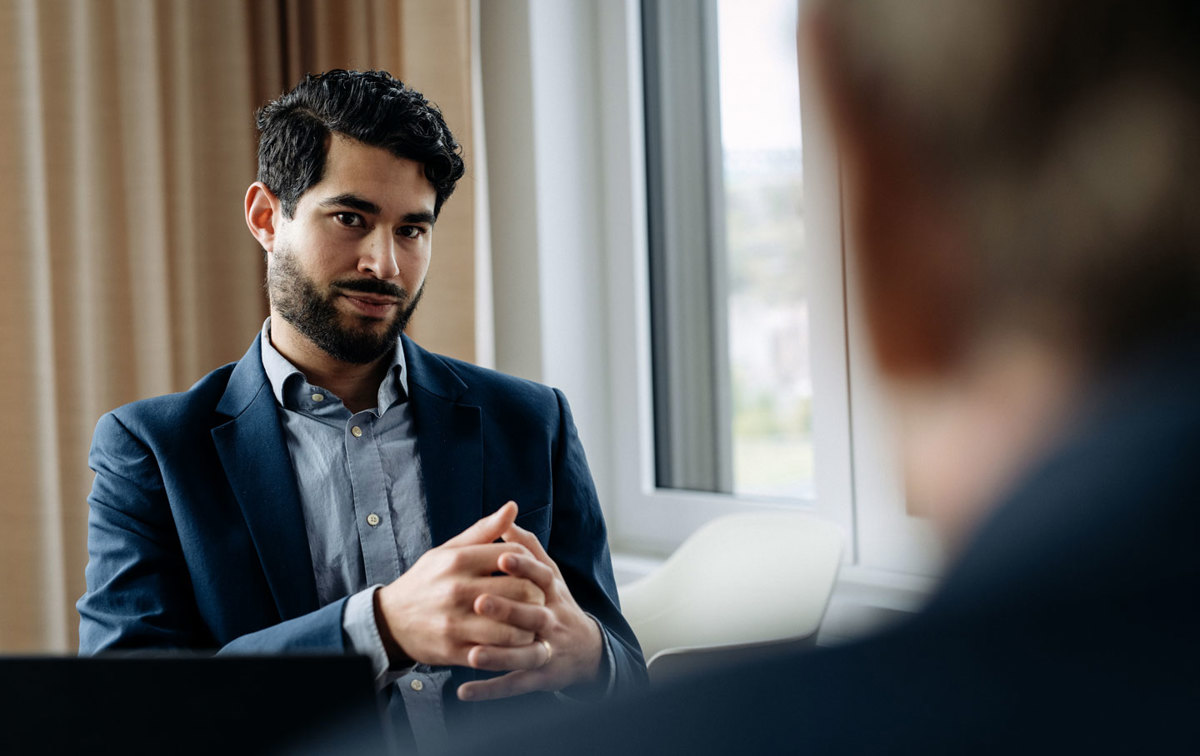 Ein Mann im Anzug hat die Hände vor sich verschränkt und spricht mit einer Person in der Unschärfe im Vordergrund