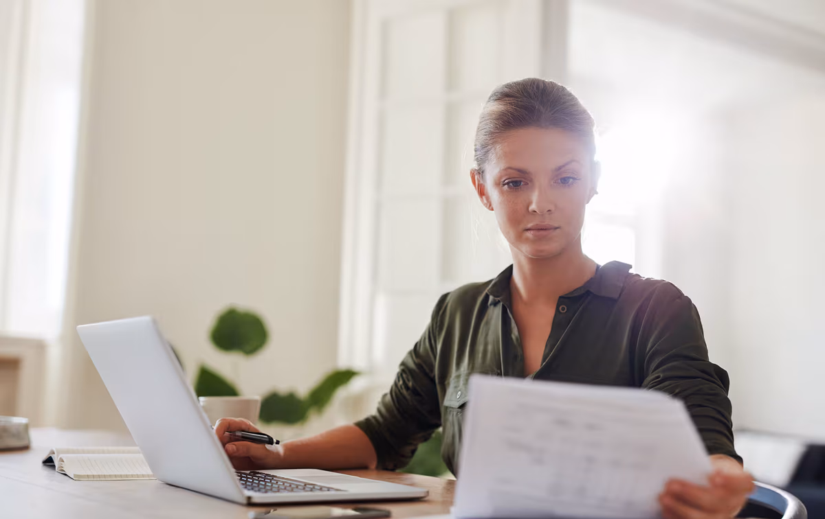 Eine Frau liest ein Dokument am Schreibtisch, während der Laptop aufgeklappt vor ihr steht.